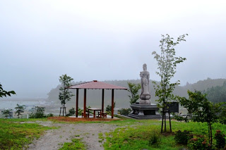 霧がかった海辺の高台に東屋と観音像が並び、周囲に若い木々が植えられた静かな広場の写真