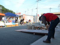 一段高くなった場所に立った男性が前屈の姿勢をしており、屋外のテントの横で男性の方を向いて立った人々が同じポーズをしている体操の様子の写真