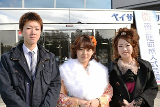 成人式の看板の前で、スーツ姿の佐藤 慶治さん、振袖と黒いコートを着た女性2名が並んで写っている写真