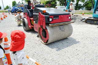 赤い帽子をかぶった園児たちが見守る中、赤いロードローラーが砂利道を転圧している様子を写した写真