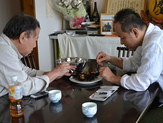 食堂のテーブルで、向かい合って座った二人の男性がウニを爪楊枝を使って食べている様子の写真