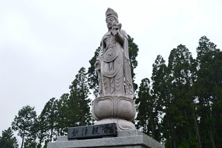 背の高い木々を背景に、蓮台に立つ優しい表情の観音像を見上げて写した写真