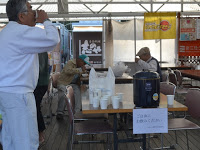 テント内のテーブルの上に紙コップと保温鍋が置かれ、その周りで数名の男性たちが飲食をしている様子の写真