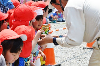 赤い帽子をかぶった園児たちが並んで立っており、作業服の男性が園児たちに手に持ったものを見せている様子の写真