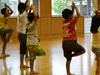 体育館の中で子供たちが両手を上にあげ手のひらを合わせ、右足を上げている同じポーズをしている様子を写した写真