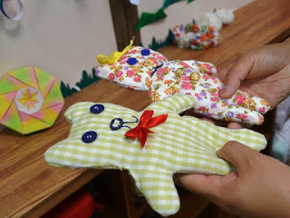 二体のクマのような手作り布人形で、一体は緑と白のチェック柄で青いボタンの目と青い刺繍の鼻と口、赤いリボンを首につけ、もう一体は花柄で青いボタンの目と刺繍の顔、黄色いリボンを頭にをつけたものを、木製の台の前で両手に持っている写真