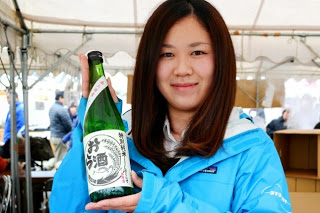 水色のジャンバーを着た女性が緑色の瓶にはいったお酒を持っている写真