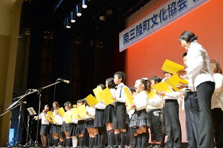 白いシャツを着た子どもたちと先生が黄色い楽譜を持ち合唱をしている様子の写真