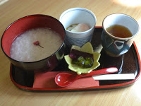 赤いお椀に入ったお粥、白い小鉢に入った温泉卵、小皿に盛られた漬物、そして湯呑みに入ったお茶が黒いお盆の上に並べられている写真