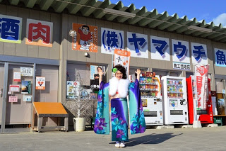 紫と青の華やかな振袖を着た女性が、自動販売機のある店舗前で両手を上げてポーズをとっている写真