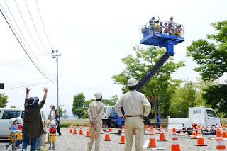 青い高所作業車のバケットに園児たちが乗っており、地上の作業員や保育士、子どもたちがそちらを見上げたり手を振ったりしている様子の写真
