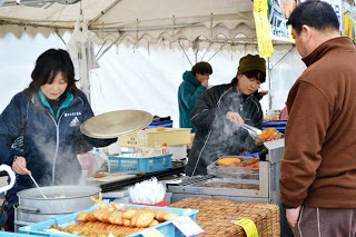 テントの下で女性たちが大きな鍋や鉄板を使って調理をしており、手前には、揚げ物や練り物（おでん風の食べ物、さつま揚げのようなもの）が並び、対面に茶色の上着を着た男性客が立っている屋台の様子を写した写真