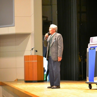 スーツ姿の男性がマイクを持ち立ちながら歌を歌っている様子を右斜めから写した写真