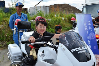 警察官が見守る中で、冠をつけた女の子が白バイにまたがって笑顔を見せている写真