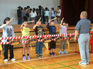 舞台前に立つ5人の児童が太鼓の前でバチを構え、目の前の指導者の合図を待ちながら練習に集中している写真