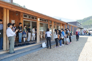 木造の建物の軒下に並んでいる人たちに、十数人の学生たちが一列に並んであいさつをしている写真