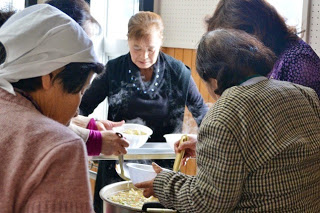 数名の女性が蒸気の立ち上る鍋から汁物をよそい、手分けして配膳している様子の写真
