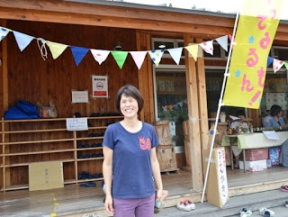 背景にはカラフルな三角旗が飾られた棚やテーブルが並んでいて、屋外イベント会場の木造の建物の前に立っている笑顔の阿部純子さんの写真