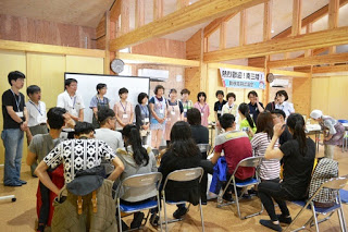 木造の室内で、前方に立つ十数人の地域住民と、イスに座った学生たちが向き合って話をしている様子の写真