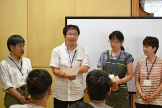 4人がホワイトボードの前に立ち、左から二番目の男性が学生たちに向けて紙を持ちながら中国語で挨拶をしている写真