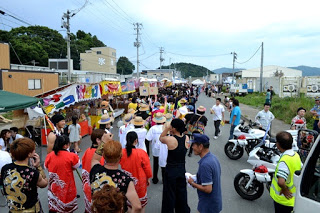 左側に屋台が並んだ奥まで続く通りを、祭りの衣装を着た人々の行列が続いている写真