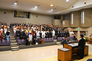 広い式典会場で、新成人たちが前方の壇上を向いて起立し、代表の新成人の男性が、壇上で関係者の男性と向かい合って立ち誓詞を読んでいる厳かな雰囲気の中で式が進行している、成人式典中の会場内の写真