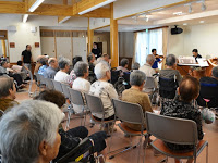 施設のホールに並んだ椅子に施設利用者の方々が座り、前方で黄緑色のTシャツを着た合唱団の歌と楽器の演奏を聴いている写真