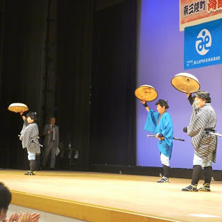 笠を上にあげた着流し姿の3名が舞台で踊りを踊っている写真