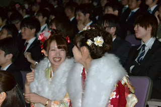 式典会場の座席に並んで座る振袖姿の女性たちが、隣同士で楽しそうに満面の笑みで会話をしている様子を写した写真