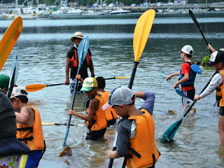 たくさんの子どもたちがライフジャケットを着用し、カヤックのパドルを手に持ちながら浅瀬で水の掻き方の練習をしている写真