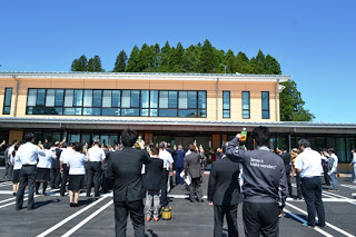 大勢の関係者が駐車場に集まり、建物に向かって手を挙げて乾杯をしている様子を背後から写した写真