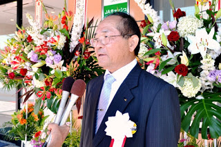 花で彩られた背景の前で、胸に白いリボンをつけた男性がインタビューを受けている写真