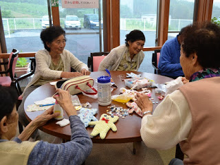 大きな窓から外の景色が見える室内で、円形のテーブルを囲んで数人の高齢の女性が座り、布やハサミ、ぬいぐるみなどの材料を使って手芸活動に取り組んでいる写真