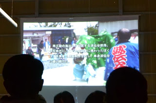 薄暗い会場内で、観客のシルエット越しに、スクリーンに地域の祭りの様子を撮影した映像が映し出されている写真