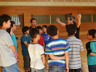 体育館の一角で男性講師が腕を広げながら児童たちに説明しており、児童が輪になって聞いている写真