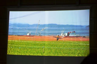 スクリーンに青々とした田んぼが広がり、その向こうに漁船が浮かぶ穏やかな海と遠くの山並みが映し出されている写真