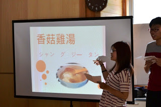 スクリーンに中国料理の名前と写真が映され、女子学生が中国料理の紹介をしている写真