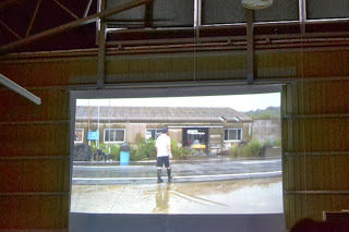 白いスクリーンに、雨が降る道路の前で白いシャツと黒い長靴を履いた人物が映っている様子の写真