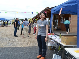 青空の下、屋外イベント会場に複数のテントが並んでいる場所に立っている笑顔の栗林美知子さんの写真