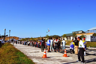 屋外の舗装された通路に整然と並び列を作った大勢の人々が待機している写真