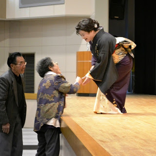 舞台の下に2名の人が並んでいて、金と黒の着物を着た女性と握手をかわしている写真