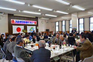 「入谷地区 新春のつどい」と書かれた横断幕と国旗と町旗が前に飾られた室内で、多くの参加者が長机を囲んで食事と会話を楽しんでいる様子の写真