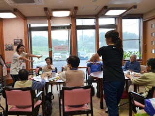 大きな窓から自然光が差し込む明るい室内で、テーブルの周りに座った高齢者の方々と、その前後に立った職員の女性2人が腕を前に伸ばして体操をしている様子の写真