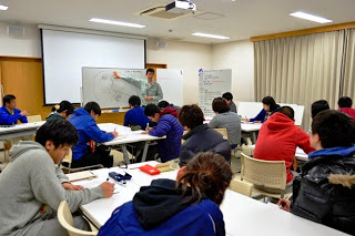 前方のホワイトボードを使い説明をしている男性の話を各班ごとに分かれた参加者がノートに記入しながら聞いている写真