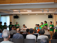 部屋の前方で緑色の服を着た人々が歌っている様子を後方から観客が見ている様子を写した写真