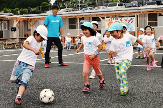 水色と白の帽子をかぶった園児たちがサッカーボールを追いかけている様子を笑顔でみている長谷部選手の写真