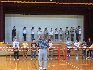 舞台上に立つ笛担当の子どもたちと、前方で太鼓を叩く子どもたちが並び、男性講師が前に立って指導している写真