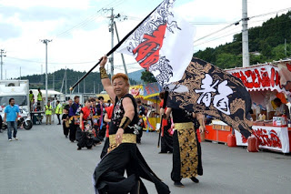 同じグループのメンバーが並んでいるなか、お揃いの衣装を着た男性が大きな旗を高く掲げて先頭に立っている写真