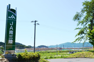 青空と復興中の広い土地が広がっている背景の手前に、「JA南三陸」と書かれた緑の看板が立っている写真
