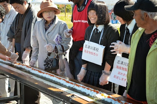 長テーブルの上に広げた巨大な巻き寿司の具材を囲んで、手袋をした人々がスタンバイしている写真